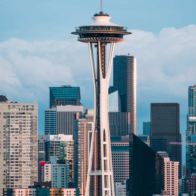 The image shows the Space Needle in Seattle with a backdrop of the city's skyline and tall buildings, under a clear sky.