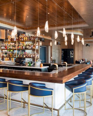 A modern bar setup with high chairs, pendant lights, a stocked bar, and seating areas in a stylish, open space.