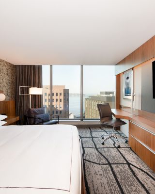 A modern hotel room with a large bed, desk, office chair, wall-mounted TV, and floor-to-ceiling windows offering a cityscape view, ending the sentence.