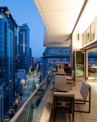 A modern cityscape view from a high-rise balcony at dusk, featuring outdoor seating and furniture with city buildings and streetlights in the background.