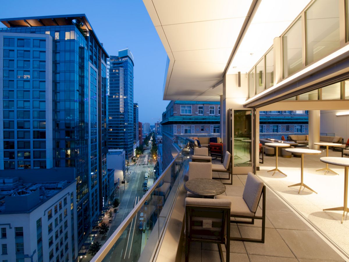 A modern cityscape with high-rise buildings is visible from a stylish rooftop terrace equipped with seating and tables.