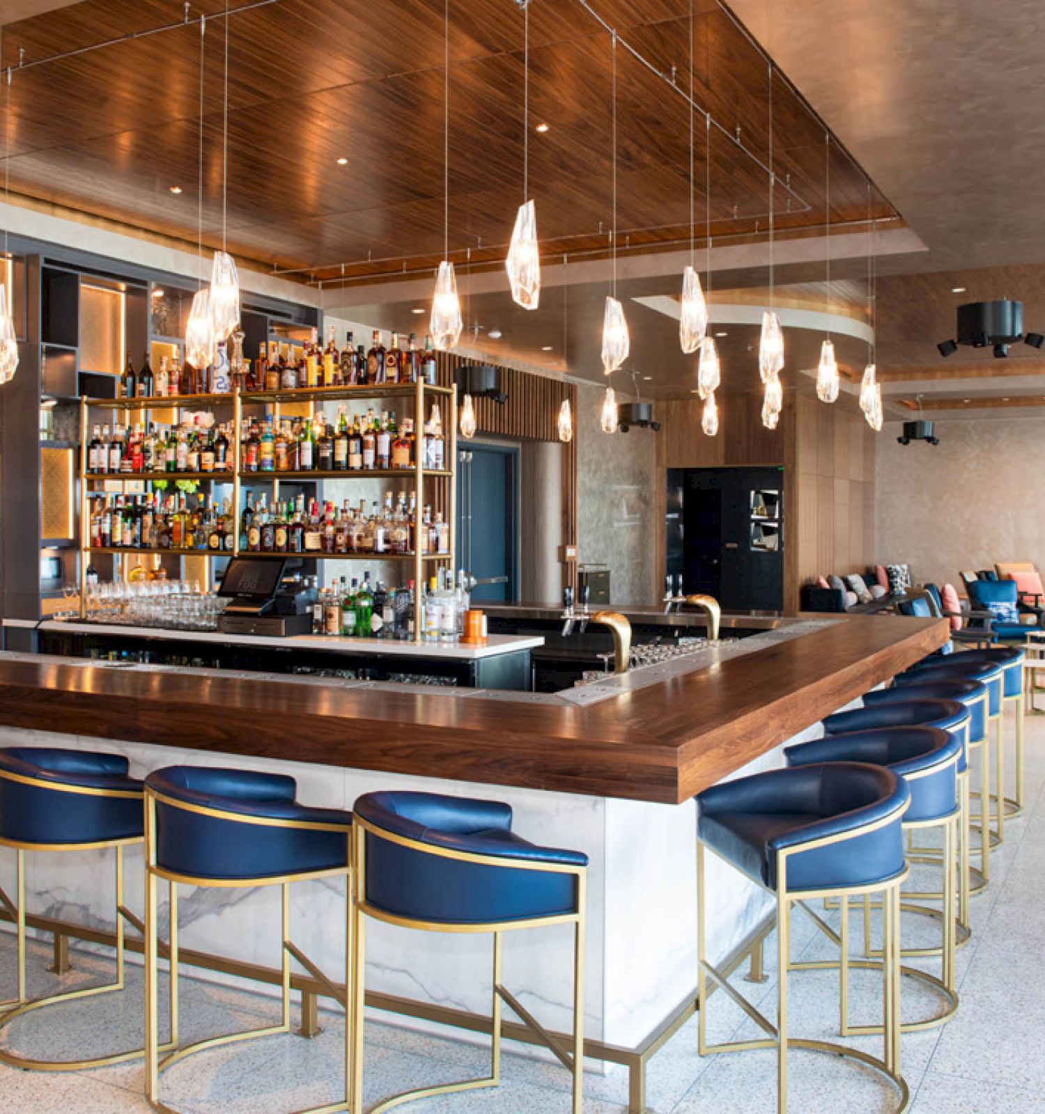 The image shows a modern bar with a wooden counter, blue barstools, hanging lights, and a well-stocked bar in a stylish, spacious setting.