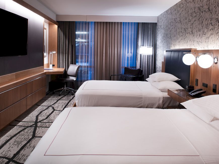 The image shows a hotel room with two neatly made beds, a wall-mounted TV, a desk with a chair, modern decor, and ample lighting from lamps and windows.