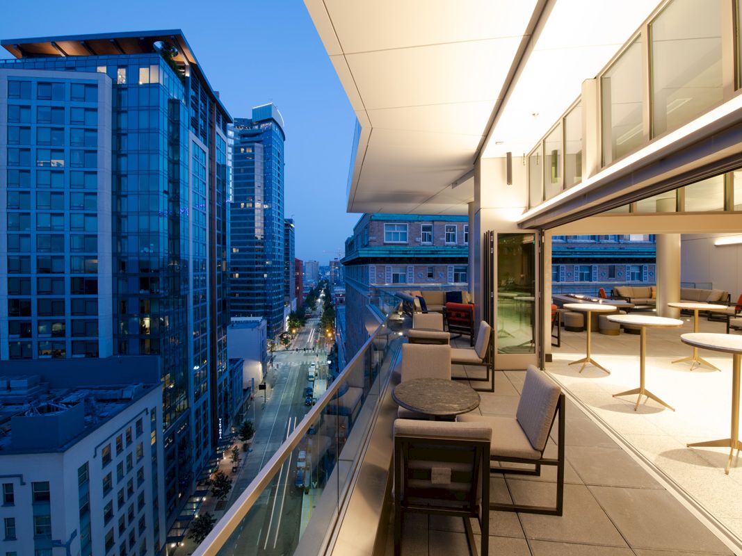 The image shows a modern rooftop terrace with tables and chairs, overlooking a city street lined with tall buildings at dusk.