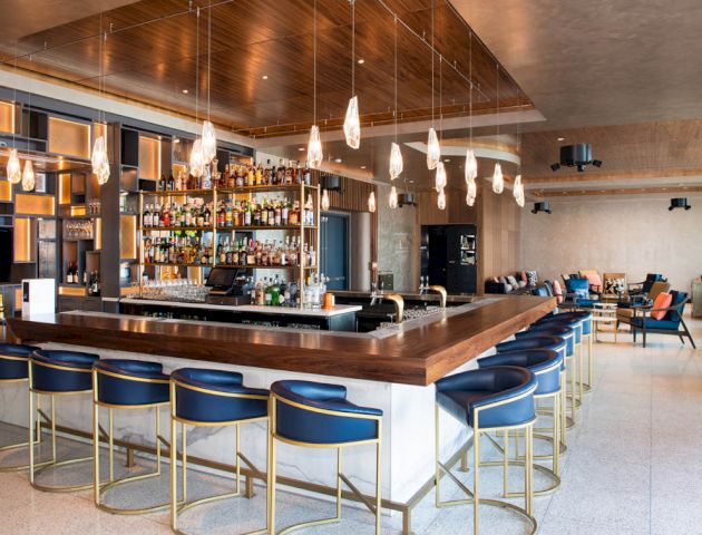 The image shows a modern bar with blue bar stools, a wooden counter, hanging light fixtures, and shelves of liquor. Lounge area in the background.