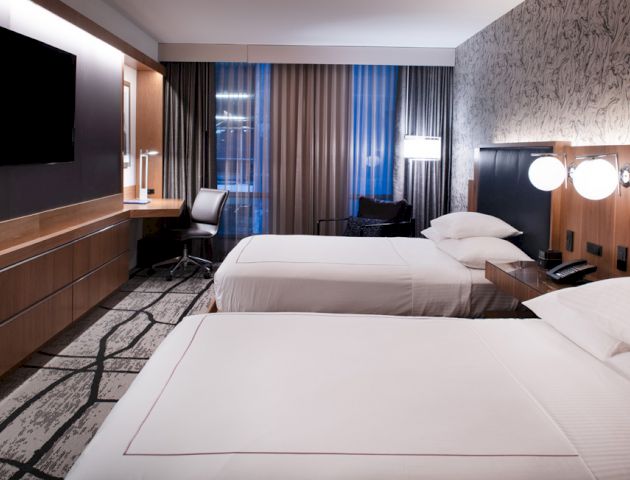 This image shows a modern hotel room with two neatly made beds, a desk, a chair, a wall-mounted TV, lamps, and stylish decor.