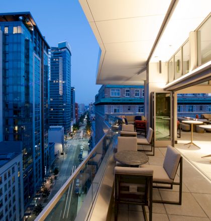 A modern cityscape at dusk with tall buildings, seen from a balcony with tables and chairs, looking down a street lined with more buildings.