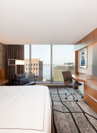 This image shows a modern hotel room with a large bed, a desk, chair, and a view of city buildings and water.