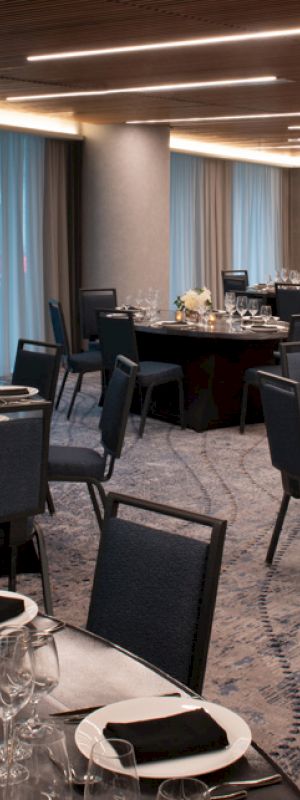 The image shows an elegantly set dining room with round tables, black chairs, folded napkins, glassware, and flower centerpieces, under modern ceiling lights.