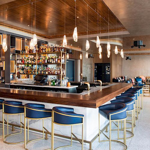 The image shows a modern bar with high stools, a well-stocked bar shelf, pendant lights, and a lounge seating area in the background.