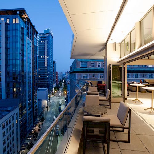 A modern balcony with tables and chairs overlooks a city street lined with tall, illuminated buildings at dusk.