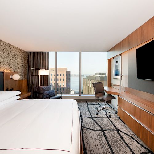 A modern hotel room with a large bed, wall-mounted TV, desk, chair, and a window with a cityscape view, featuring contemporary decor.