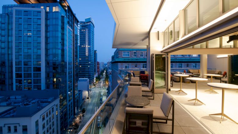 A modern balcony at dusk with tables and chairs overlooks a city street lined with tall, glass buildings.