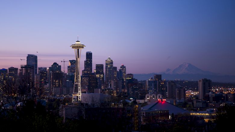 A nighttime view of the Seattle skyline, featuring the Space Needle prominently and Mount Rainier in the background, under a twilight sky.