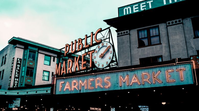 The image shows a neon sign for a "Public Market" and "Farmers Market" with a clock above. The market is in a bustling urban area with nearby shops.