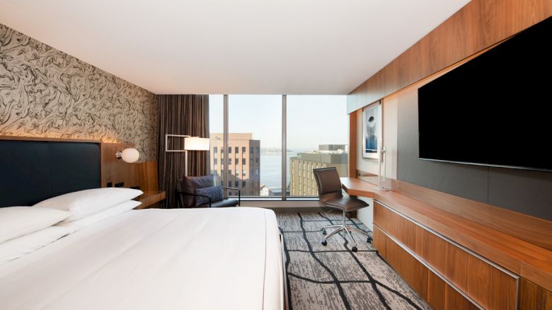 This image shows a modern hotel room with a large bed, a desk with a chair, a seating area by the window, and a large flat-screen TV.