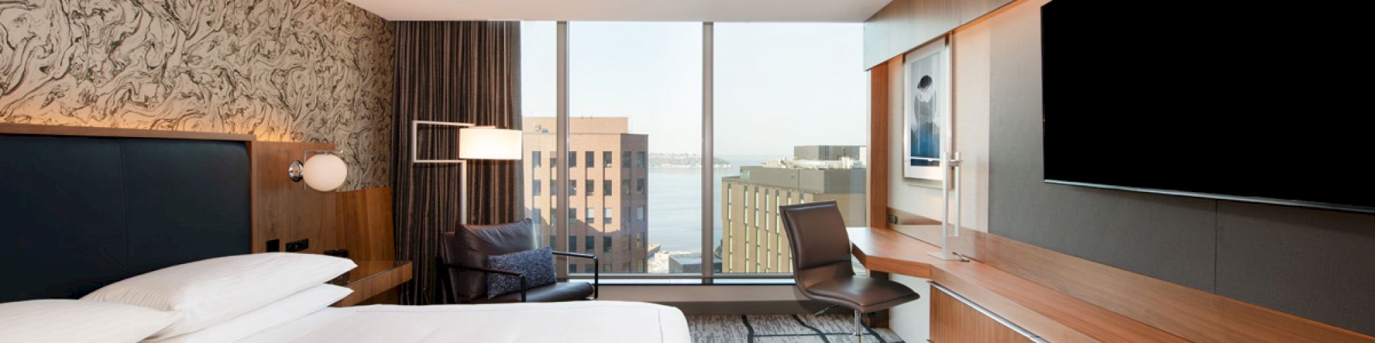 This image shows a modern hotel room with a large bed, desk, chair, and a wall-mounted TV, with a view of city buildings through a window.