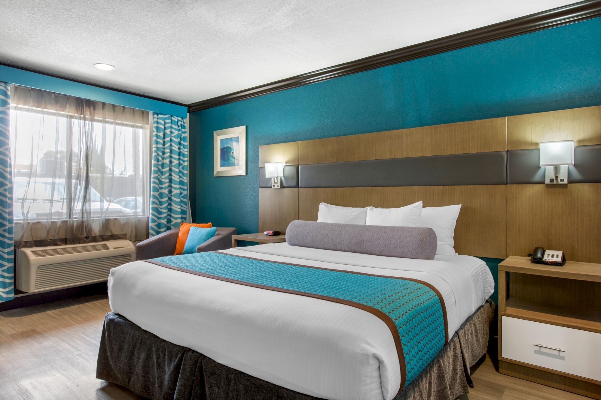 A modern hotel room with a neatly made bed, vibrant turquoise accents, bedside tables, a window with curtains, and wall-mounted lights.