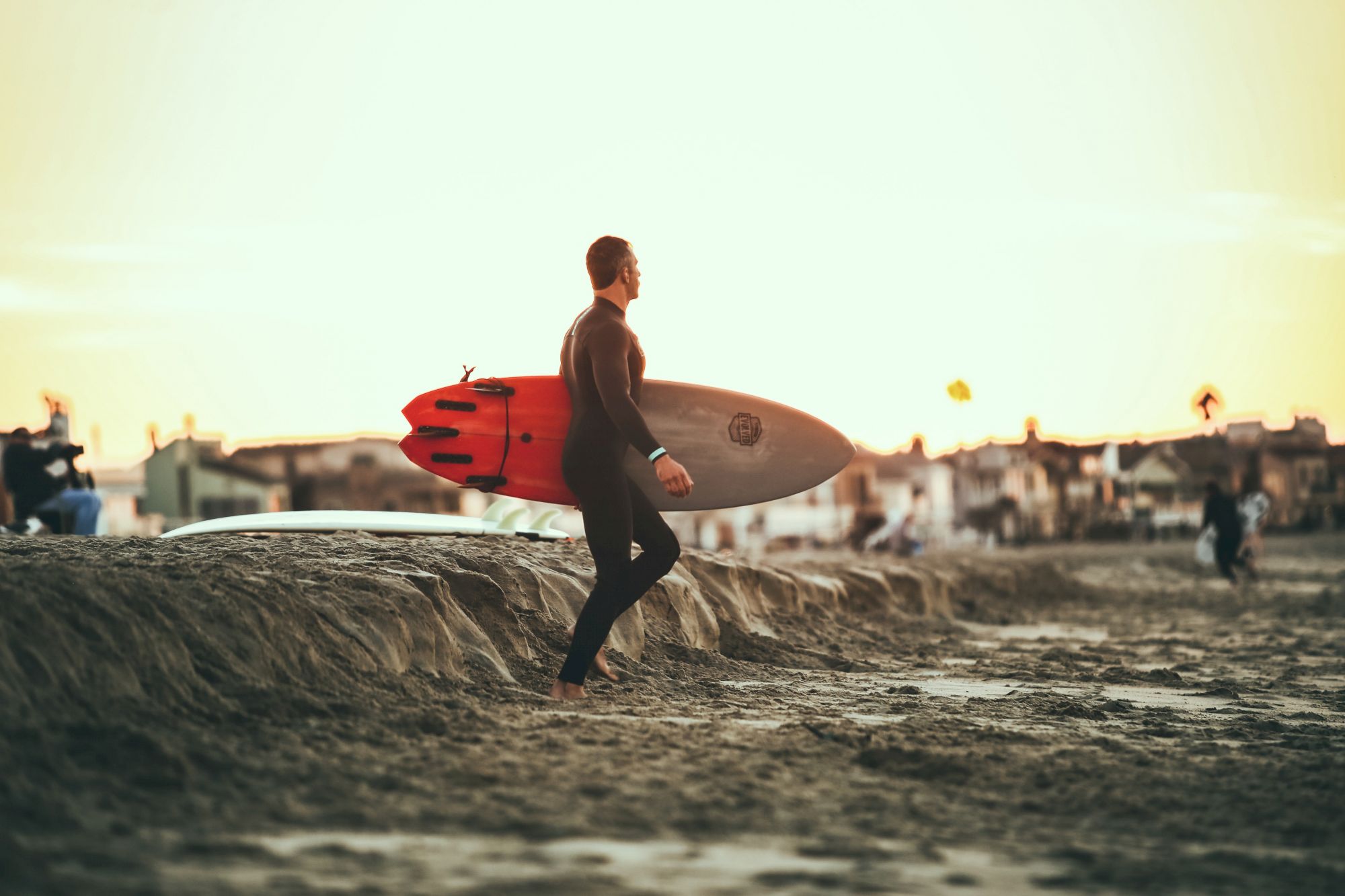 A person is holding a surfboard on a sandy beach, facing the ocean. The sun is setting, and there are buildings and a few more people in the background.