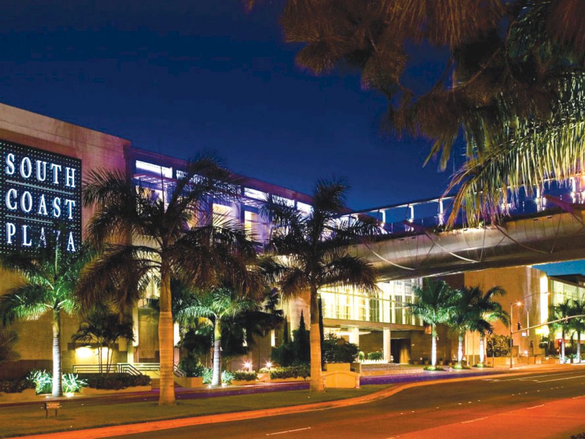 This image shows the South Coast Plaza shopping center at night, featuring illuminated signage, palm trees, and a lit pedestrian bridge over the street.