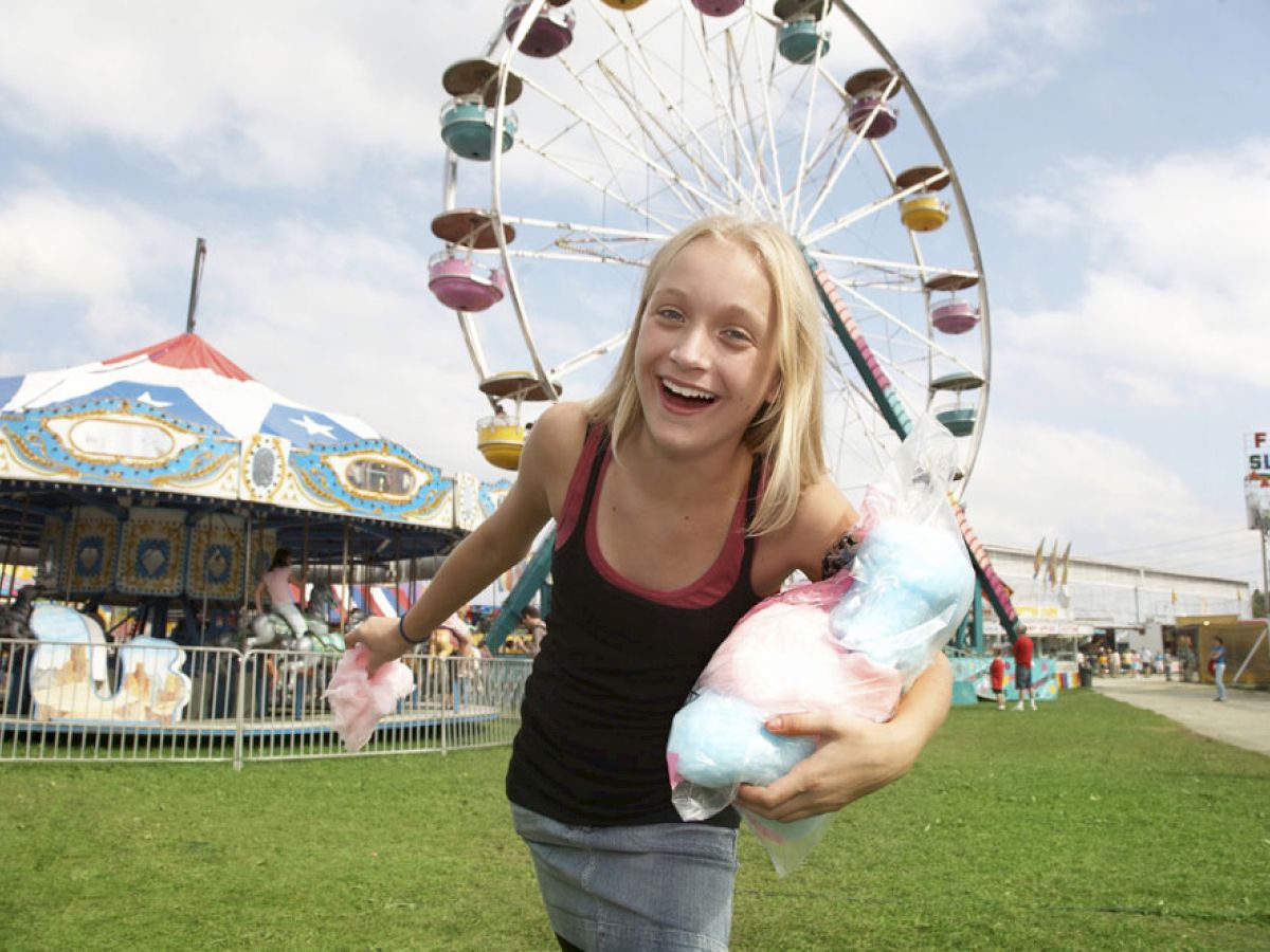 A person is enjoying a day at an amusement park, holding cotton candy with a Ferris wheel and a carousel in the background.