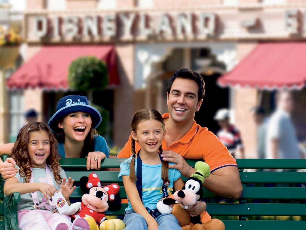 A family of four, two adults and two children, smiling and sitting on a bench at Disneyland, holding plush toys of Mickey and Goofy.