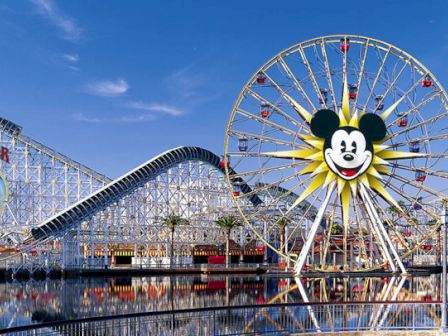 The image shows a theme park with a Ferris wheel featuring a sun and character face, roller coasters, and other amusement rides.