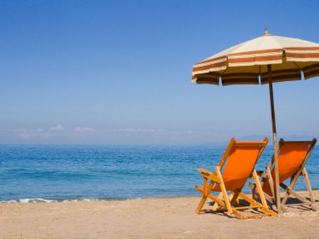 Two beach chairs under a striped umbrella are set up on a sandy beach, overlooking a calm blue ocean under a clear sky.