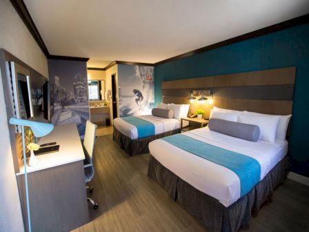 The image shows a modern hotel room with two beds, a desk with a lamp and chair, and a mural of a surfer on the wall near the bathroom.