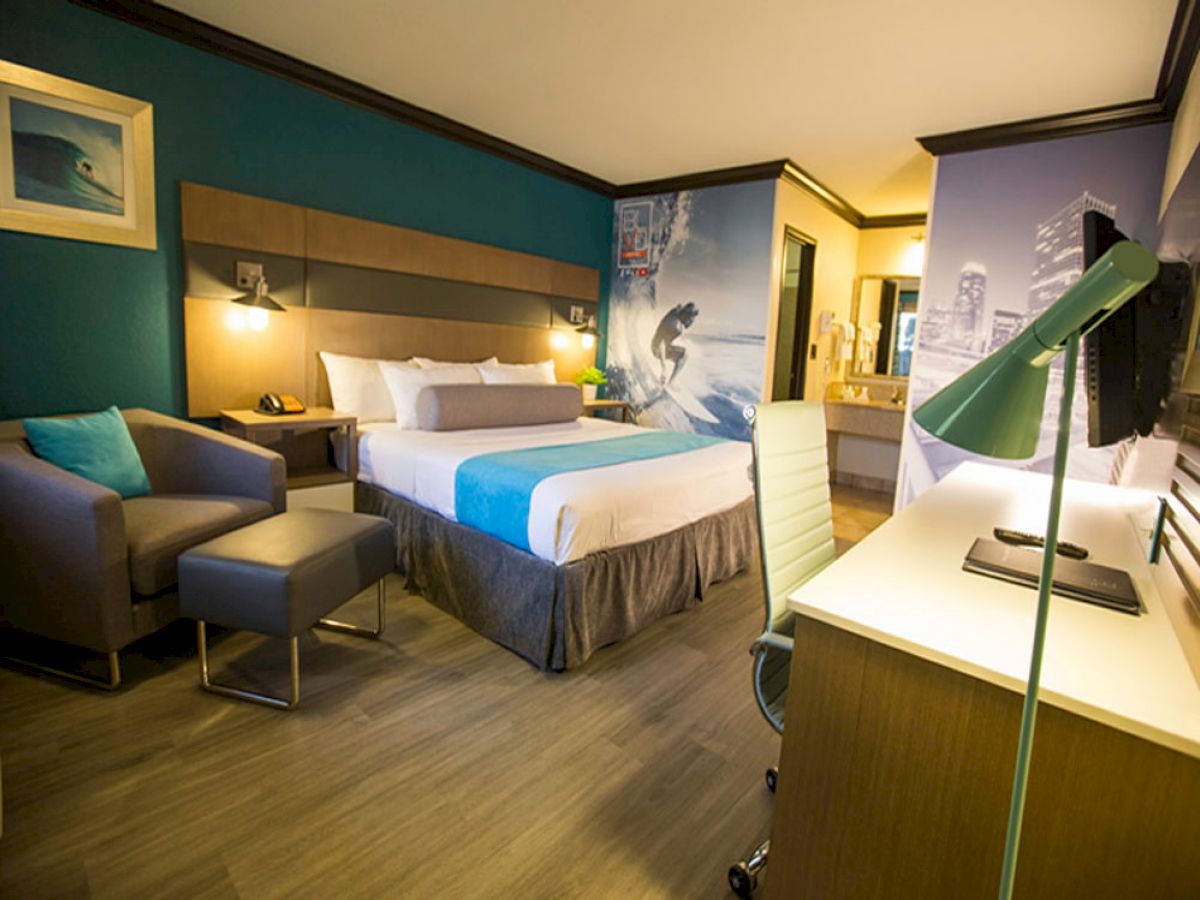 A modern hotel room with a king-sized bed, armchair, desk area, wall art featuring a surfer, and teal accents throughout the decor.