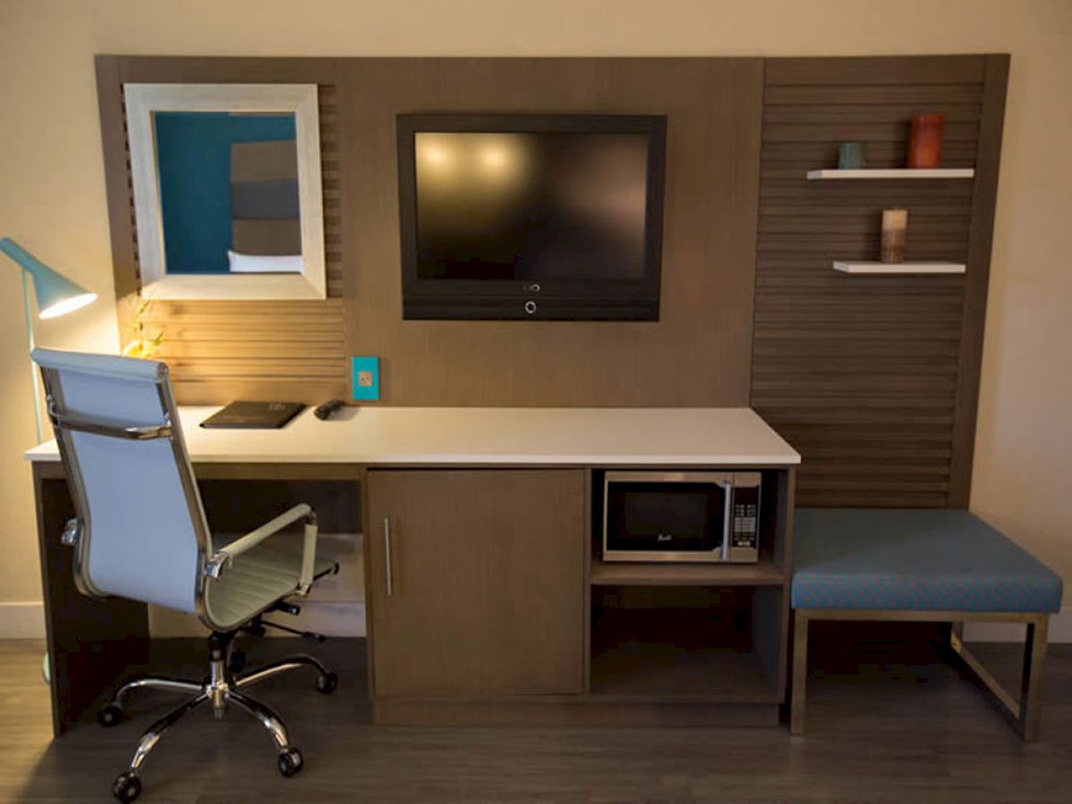 A modern workstation with a desk, chair, TV, and shelves. There's a microwave, a lamp, and a small cushion seat next to the desk, ending the sentence.