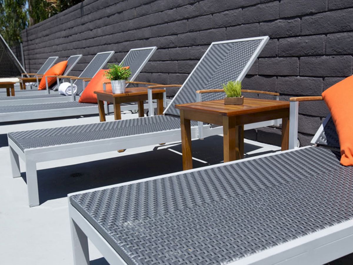 A row of outdoor lounge chairs with orange pillows and small tables between them, set against a dark brick wall.