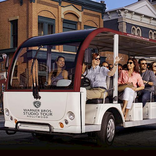 A group of people is taking a Warner Bros. Studio Tour, riding on an open-sided vehicle, passing by buildings in what appears to be a movie set.