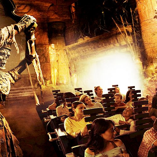 The image shows a themed amusement park ride with passengers looking at a large skeleton warrior holding a weapon in an ancient setting.