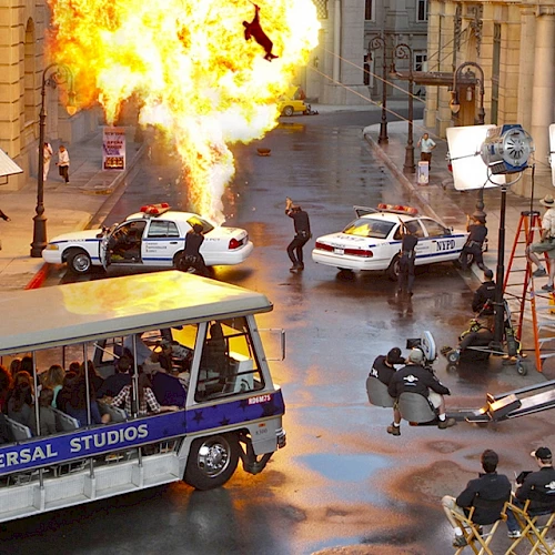 A film production set with an explosion, police cars, crew, and cameras. People on a Universal Studios tour tram watch the action.