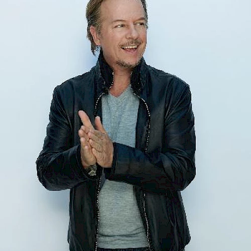A man with light hair wearing a black leather jacket and grey shirt, claps his hands and smiles against a plain background.