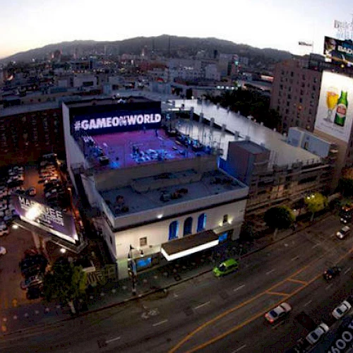 This image shows an urban cityscape at dusk with a large building prominently displaying "#GAMEONWORLD." Streets are lit, and cars are parked.