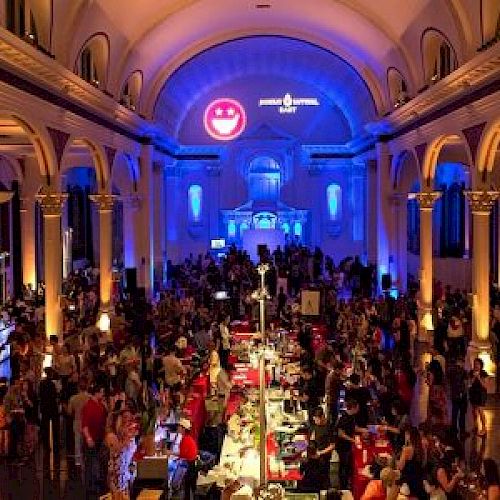 The image shows a large indoor event in a grand hall with high ceilings, arches, and columns, featuring numerous attendees and decorated with lights.