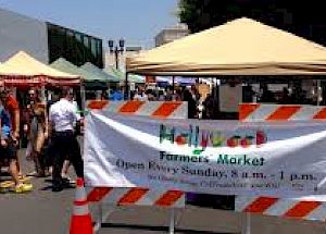 The image shows a busy farmers market with multiple stalls covered by tents. A sign reads "Hollywood Farmers' Market. Open Every Sunday, 8 a.m. - 1 p.m."