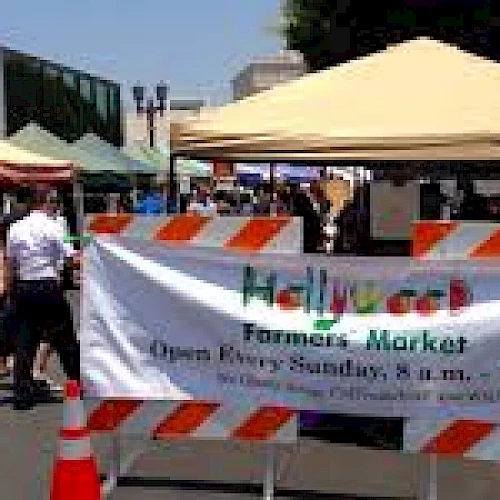 The image shows a busy farmers market with multiple stalls covered by tents. A sign reads "Hollywood Farmers' Market. Open Every Sunday, 8 a.m. - 1 p.m."