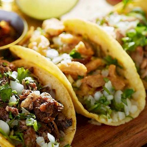 Three tacos filled with meat, onions, and cilantro on a wooden board, accompanied by a small dish of salsa in the background.